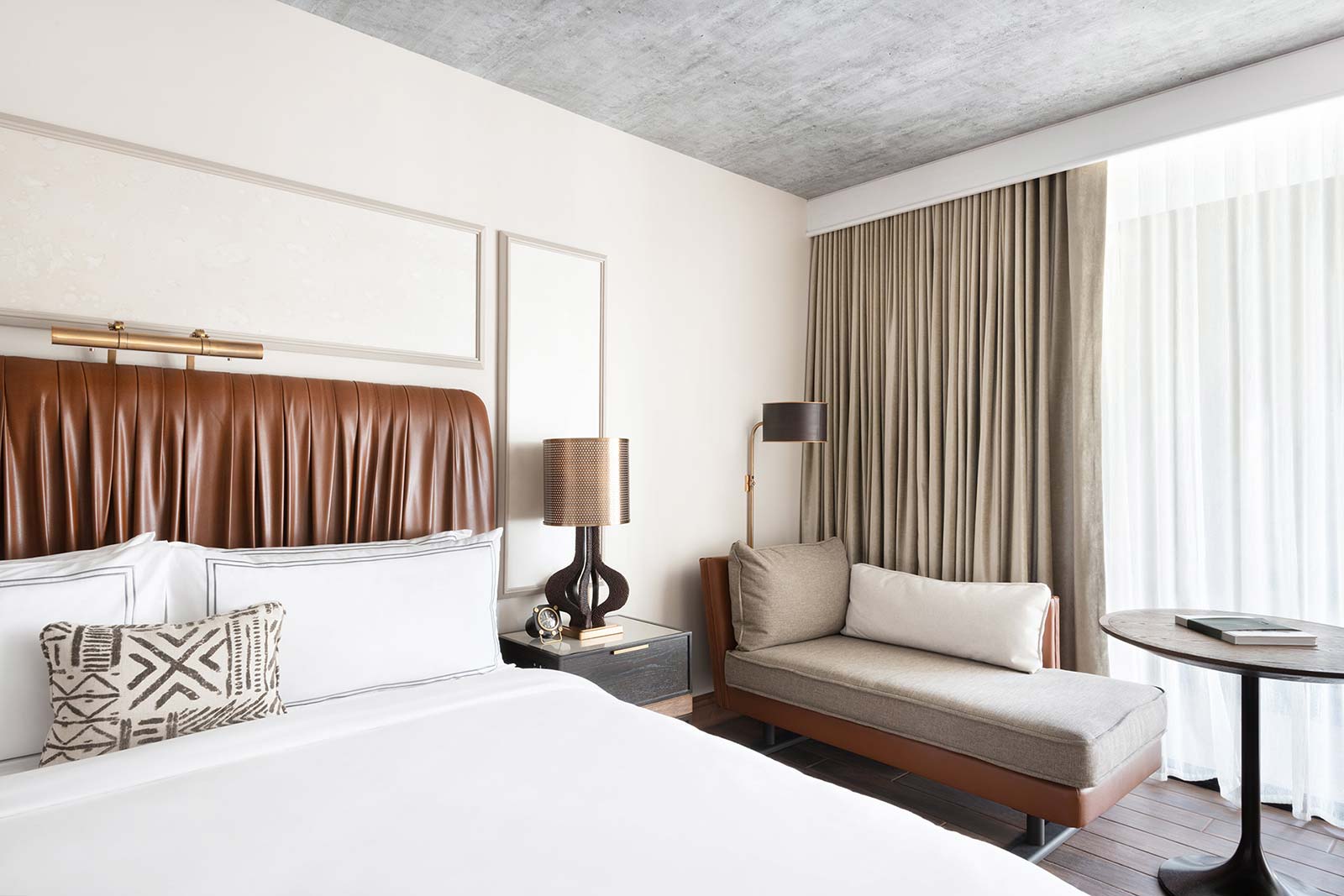 A guest room at The Ben with a leather headboard, crisp white linens, and a daybed beside a curtained window.