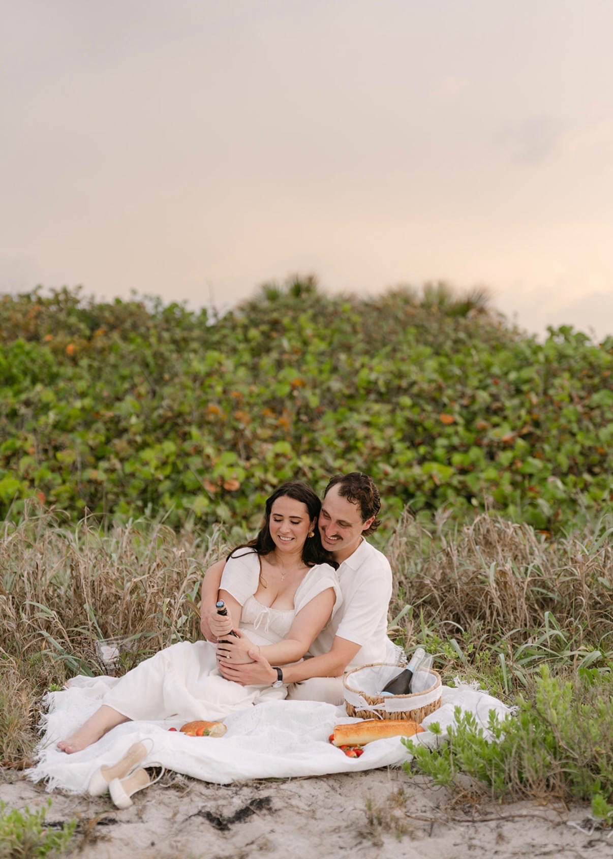 The couple sitting together on a beach picnic blanket.