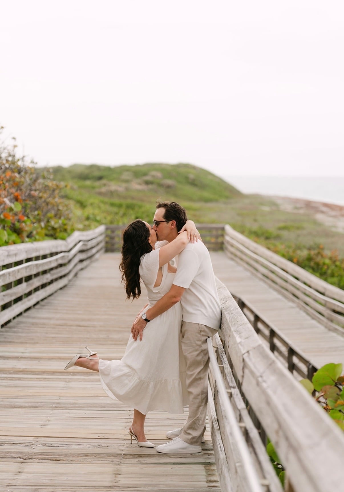 Claudia and Marcus kissing on a wooden boardwalk near the beach.
