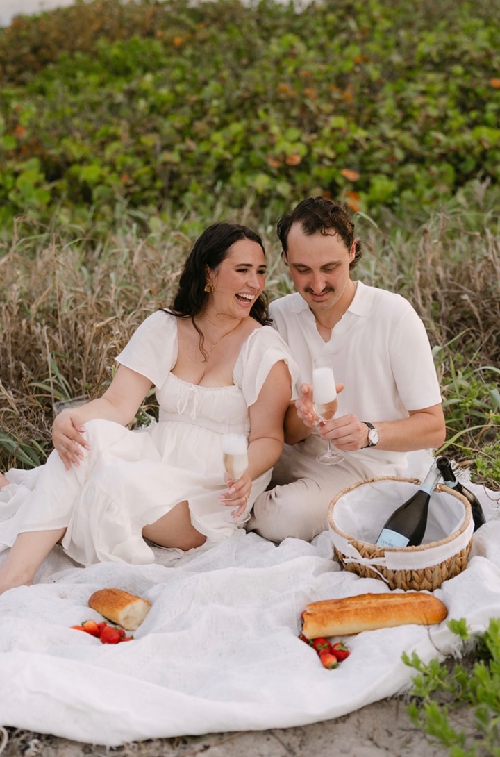 The couple laughing together over champagne on a beach picnic.