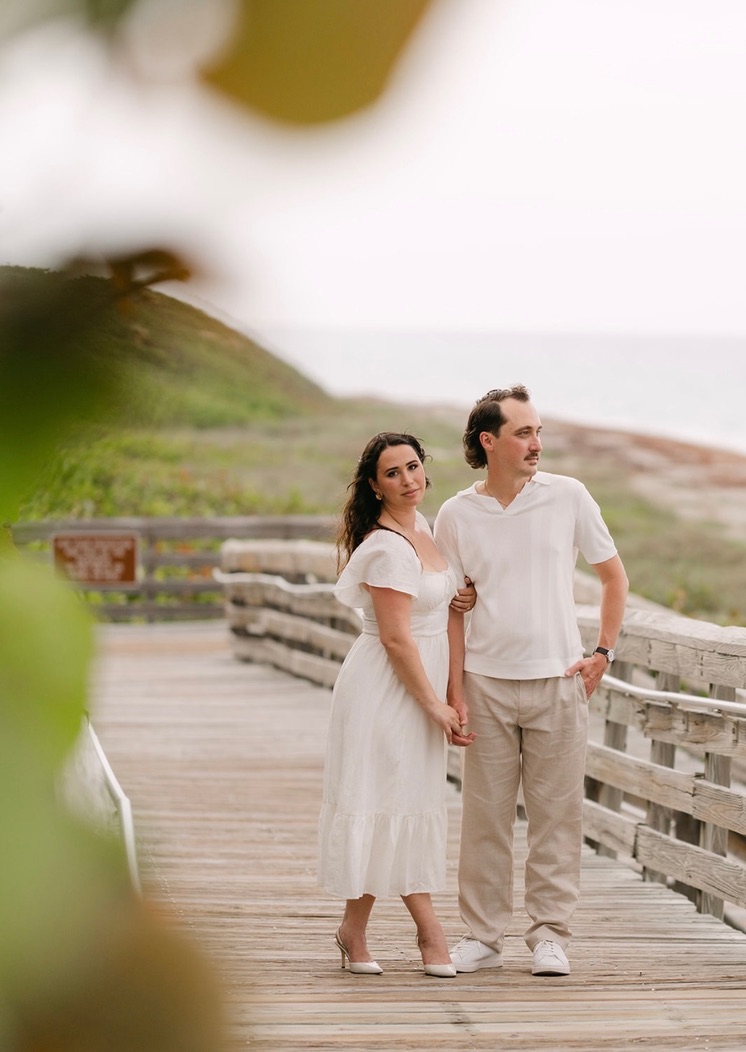 The couple holding hands on a boardwalk with beach foliage nearby.